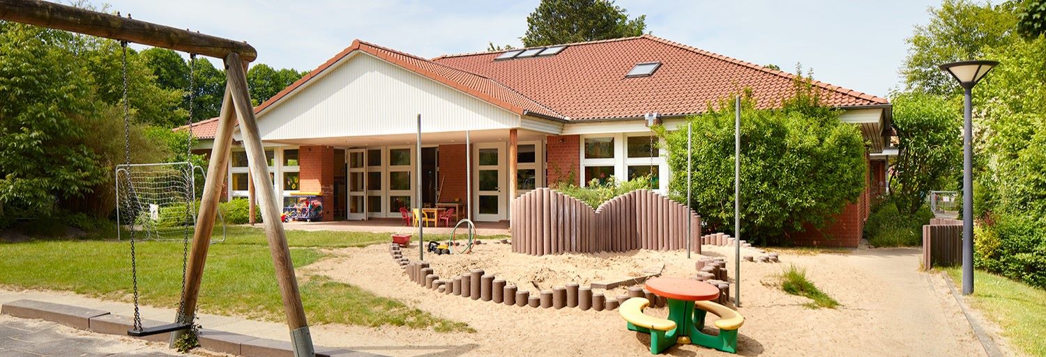 Kitagebäude mit rotem Kacheldach, ein Sand und Grasgarten ist vor dem Eingang.