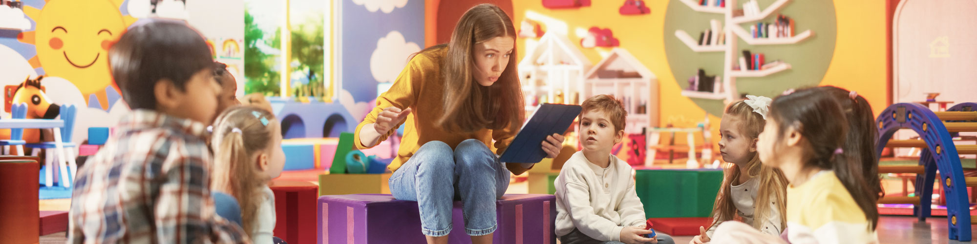Eine Erzieherin sitzt im Stuhlkreis in der Kita im bunten Gruppenraum und liest den Kindern vor.
