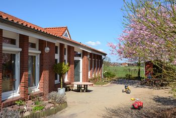 Backsteinfarbiges Kitagebäude mit Kacheldach mit einer gepflasterter Auffahrt auf der Kinderfahrzeuge stehen.