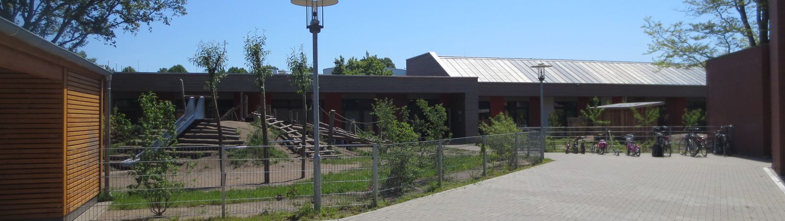 Eingezäunter Spielplatz mit kleinen Berg und Rutsche, daneben eine Auffahrt in Richtung des Kita Gebäudes.